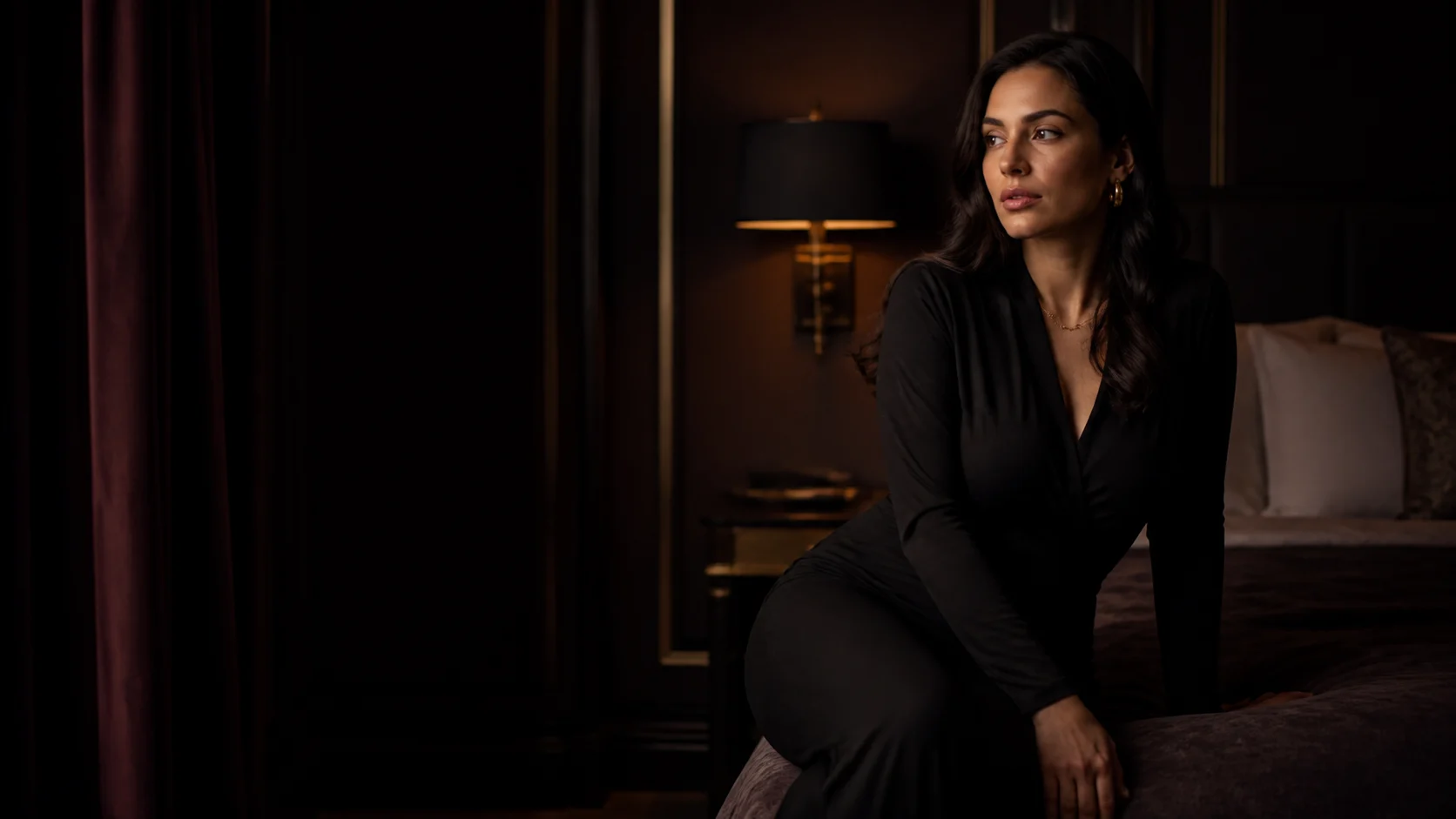 An adult woman sits in a dark plum-lit hotel room in a discreet after-hours portrait.