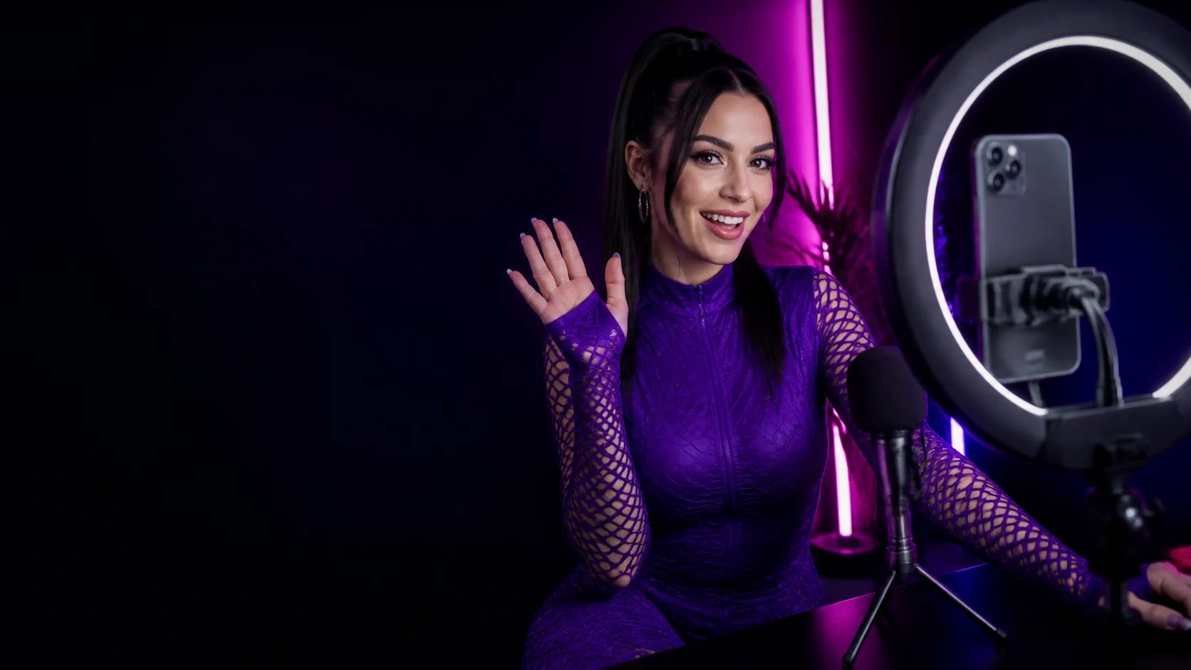An adult creator waves from a purple-neon webcam desk with ring light and microphone.