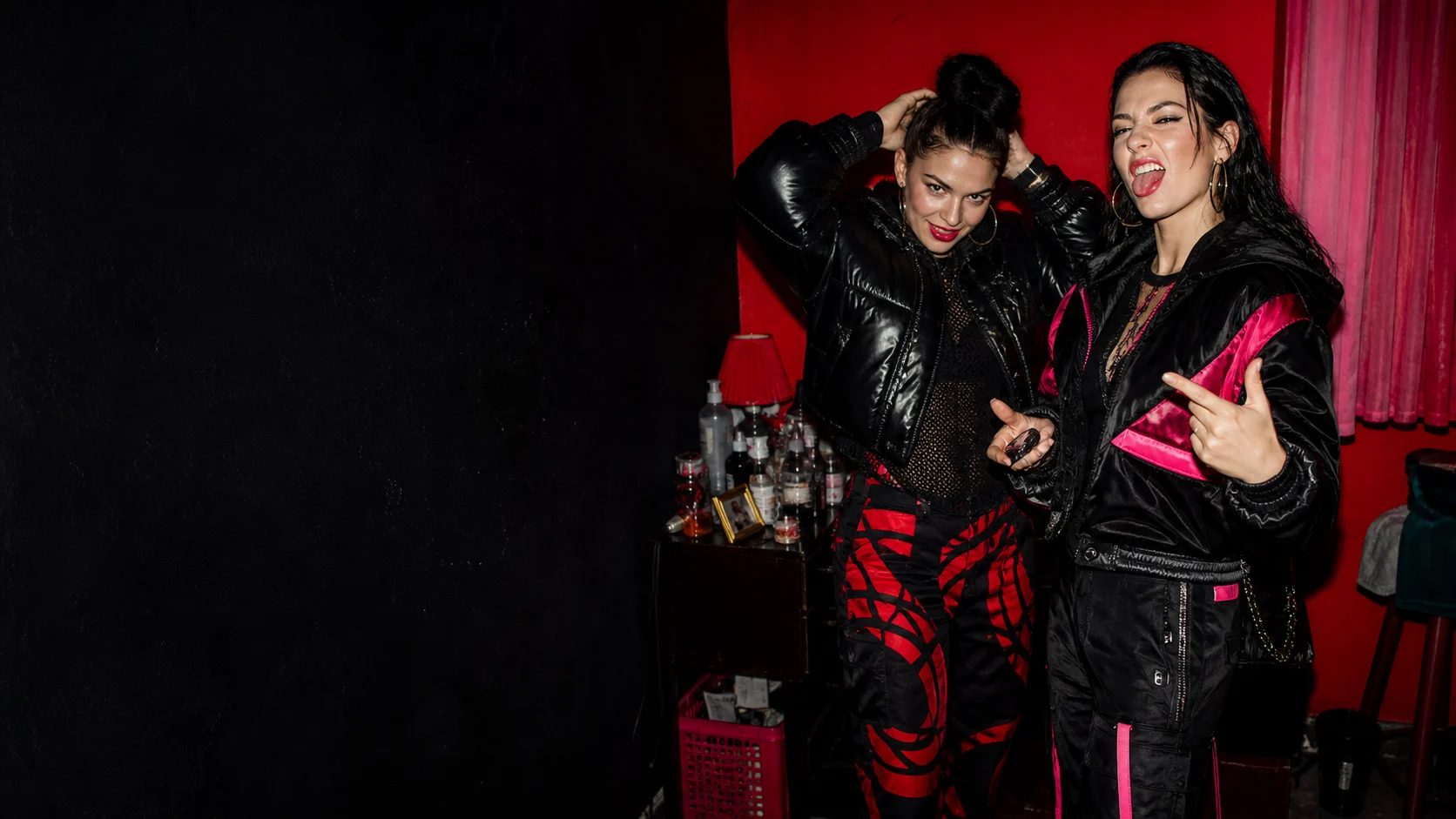 Two adult women get ready for a loud night out in a cramped red-neon apartment.
