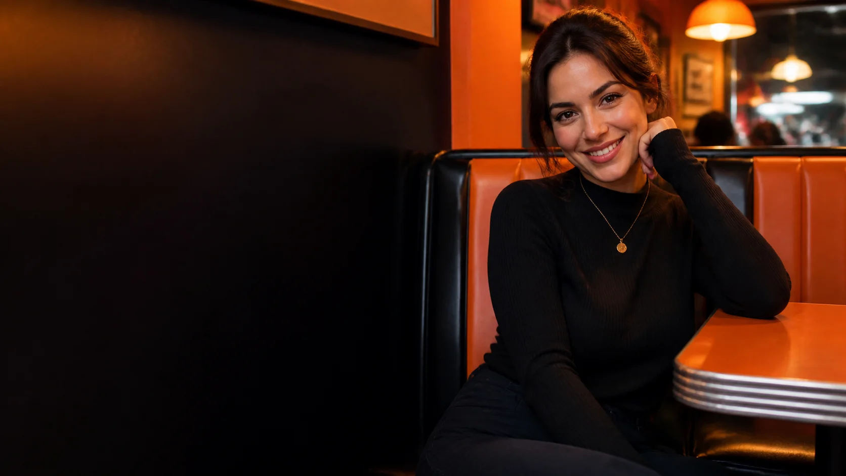 An adult woman smiles from an orange-lit late-night diner booth.